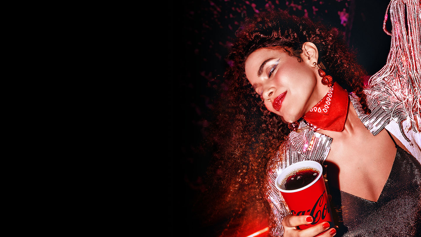 Mujer disfrutando de una bebida Coca-Cola en un evento nocturno, luciendo un look brillante, labios rojos y una bandana roja, en un ambiente festivo y lleno de energía.