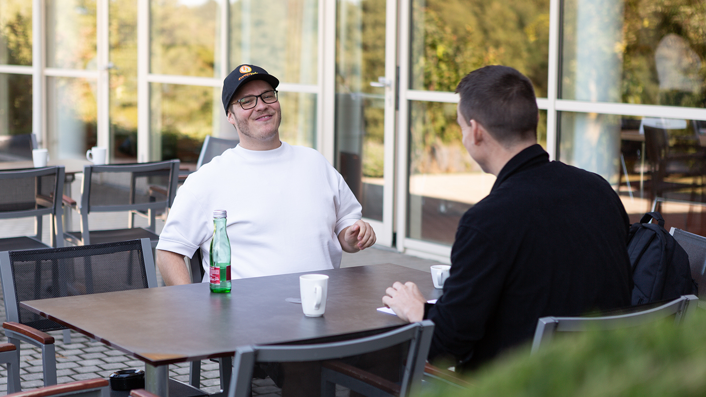 Zwei Männer sitzen draußen an einem Tisch vor großen Glasfenstern. Einer trägt ein weißes Shirt und eine schwarze Kappe, sie unterhalten sich bei Getränken in lockerer Stimmung.