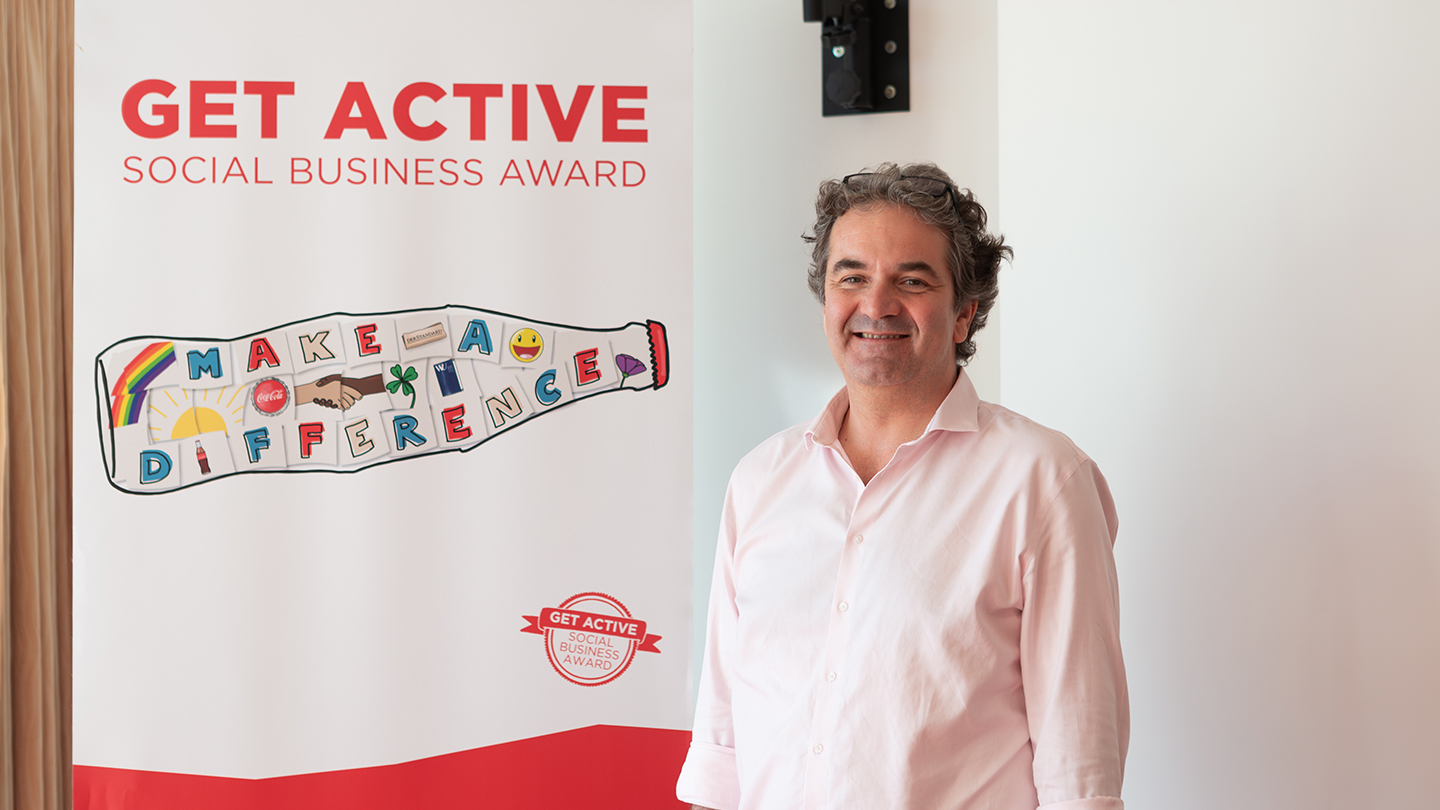 Ein Mann im rosa Hemd steht neben einem „Get active Social Business Award“-Banner mit dem Slogan „Make a Difference“. Er lächelt in die Kamera.
