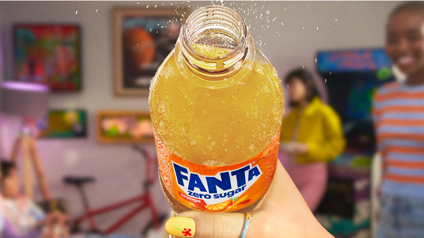 Gros plan d'une main tenant une bouteille ouverte de Fanta Orange sans sucre, montrant la boisson pétillante avec des bulles et des gouttelettes en suspension. En arrière-plan flou, on distingue une pièce colorée avec des tableaux accrochés au mur, des meubles et des personnes discutant.