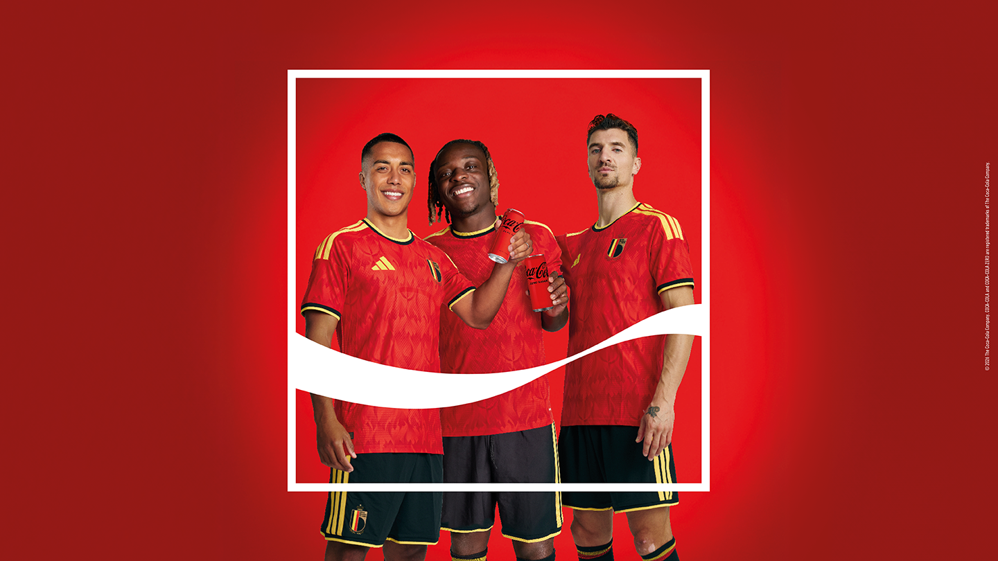 Image promotionnelle sur fond rouge représentant trois joueurs portant des maillots de football rouges, debout côte à côte dans un cadre blanc. L’un d’eux tient une bouteille Coca‑Cola rouge, et une vague blanche emblématique de la marque traverse le visuel.