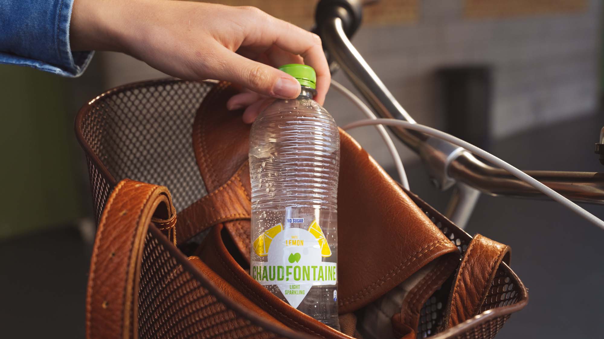 Main sortant une bouteille en plastique d’eau minérale Chaudfontaine Light Sparkling goût citron, sans sucre, avec bouchon vert, d’un sac en cuir marron posé dans un panier de vélo.