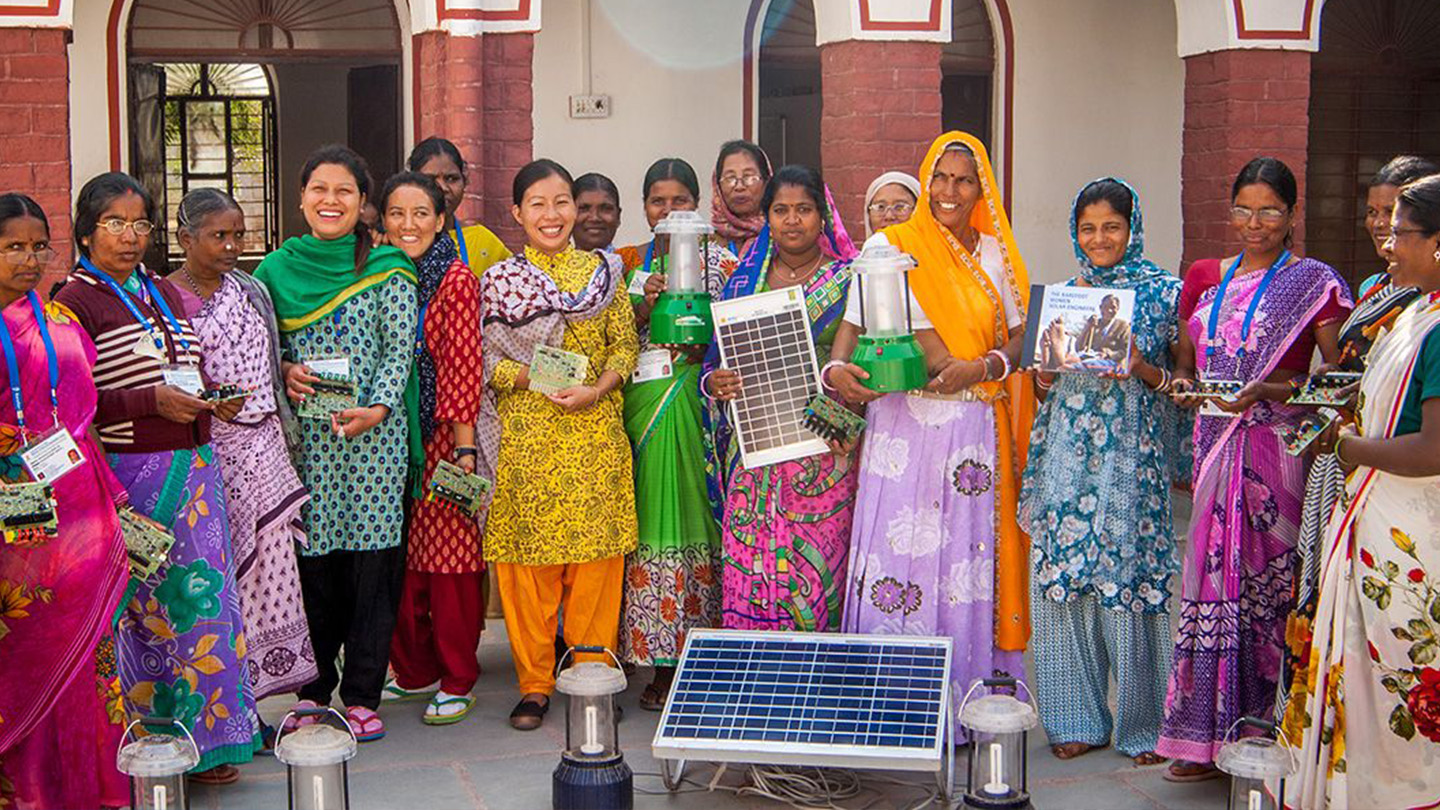 Un groupe de femmes portant des saris colorés se tient ensemble, certaines tenant des lampes solaires, des panneaux photovoltaïques ou des brochures, dans un cadre extérieur ensoleillé.