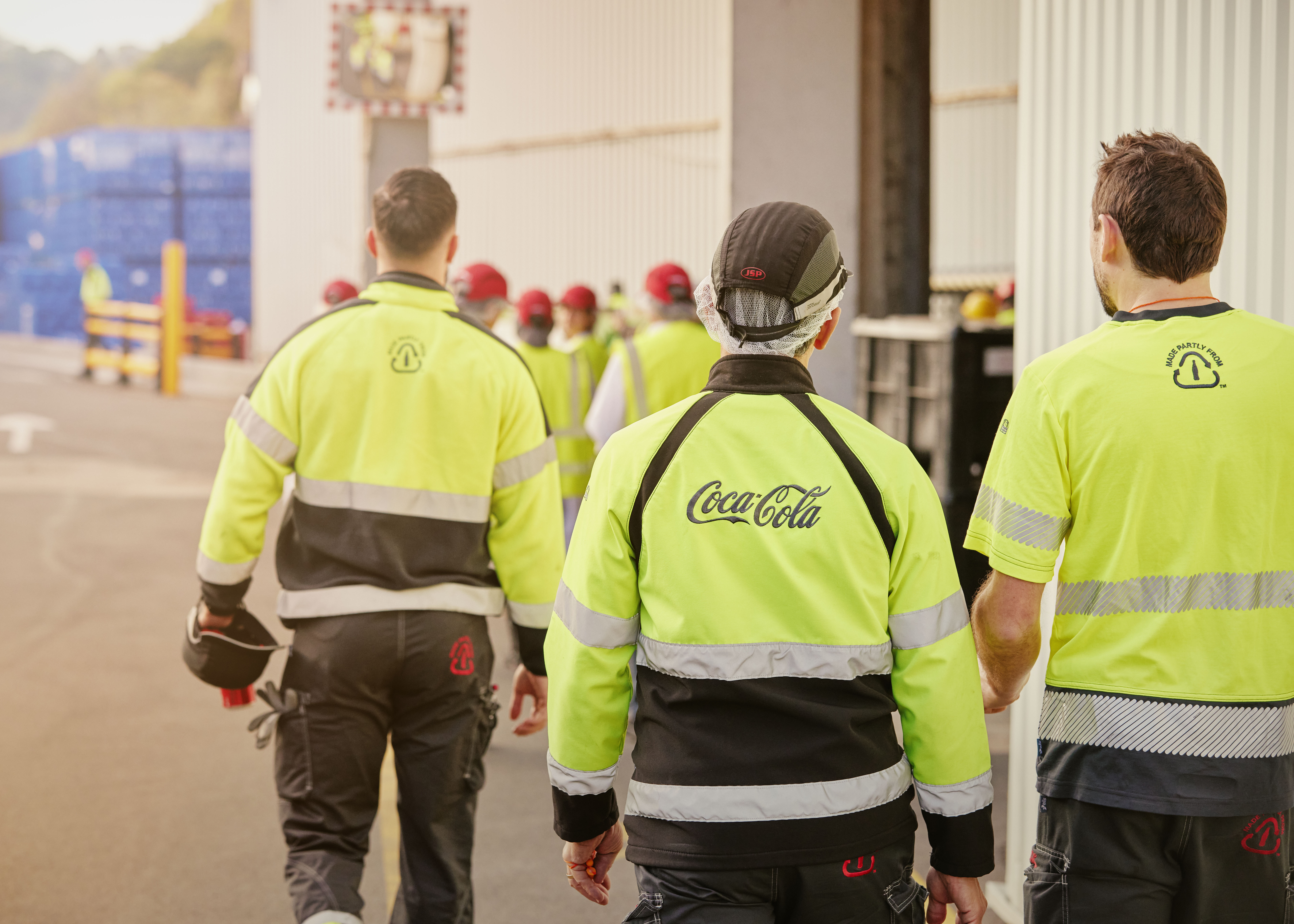 Foto met werknemers van Coca‑Cola in hoogzichtbare veiligheidskleding die naar een werkzone in de fabriek wandelen.