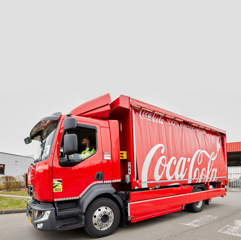 Foto met een rode 100% elektrische Coca‑Cola vrachtwagen, geparkeerd bij een industrieel gebouw.