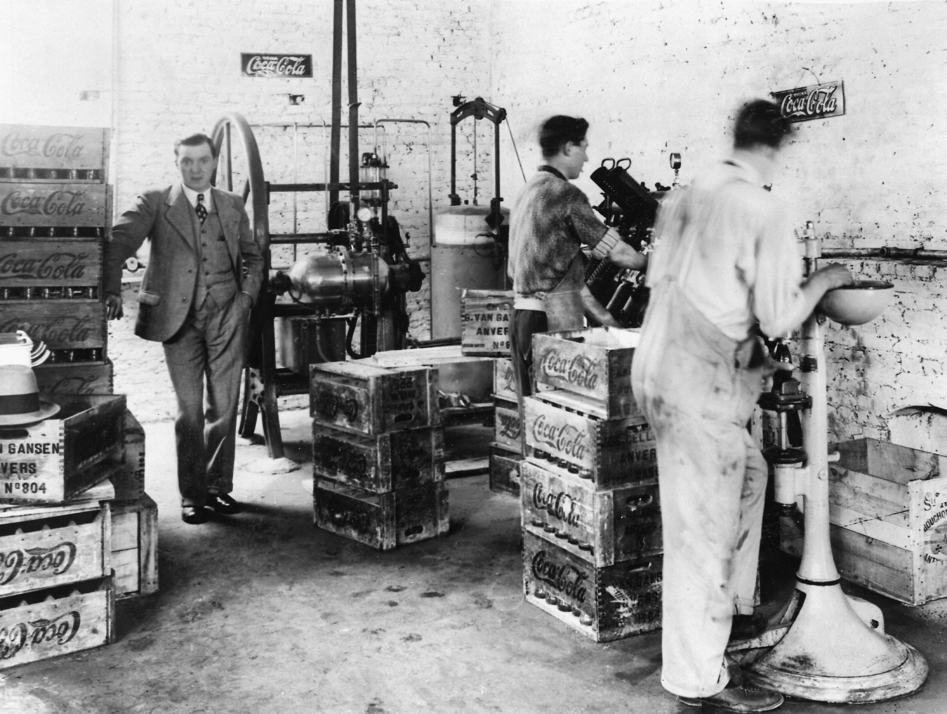 Oude foto van Coca-Cola bottelarij in Antwerpen - gustave van gansen : Foto met een oude Coca‑Cola fabriek waar mensen werken tussen houten kratten en productiemachines