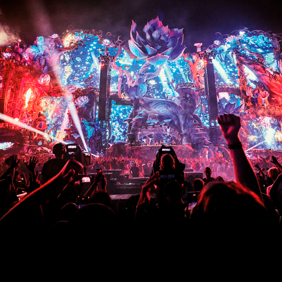 Een groots en kleurrijk muziekfestivalpodium met fantasierijke decoraties, felle lichten en visuele effecten, gezien vanaf het publiek. Op de voorgrond steken mensen hun armen en telefoons omhoog terwijl ze het optreden filmen.