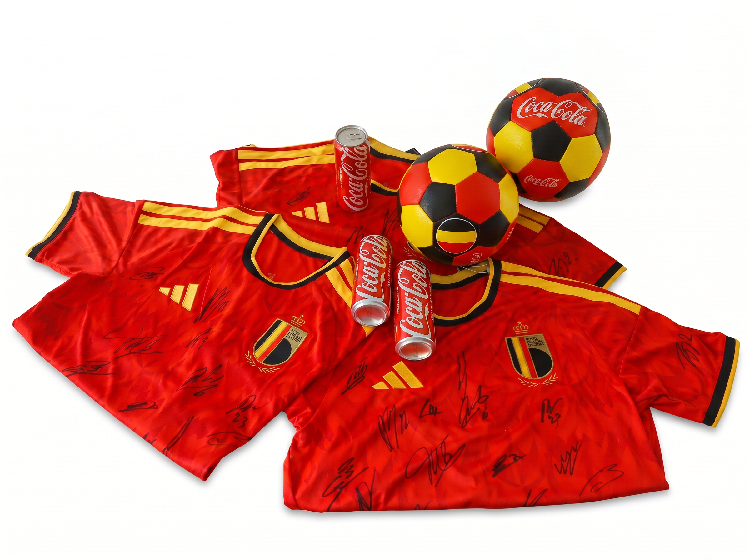 Drie rode Belgische voetbalshirts met handtekeningen liggen naast elkaar op een witte achtergrond, met meerdere Coca‑Cola blikjes en twee voetballen in Belgische kleuren erboven.