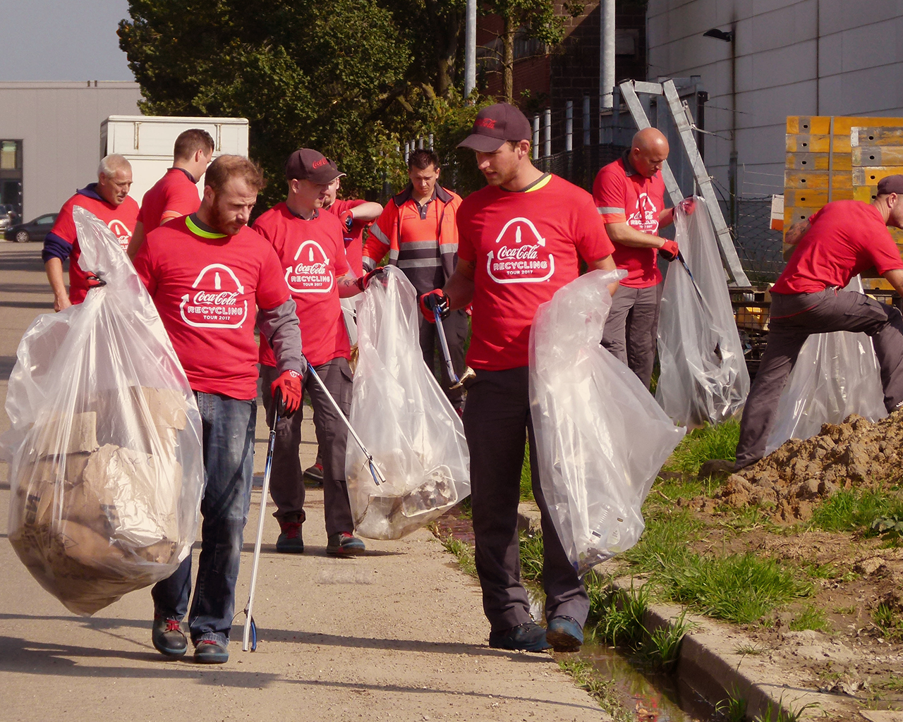 De foto toont een groep mensen in rode T-shirts met het Coca-Cola-logo en de tekst "I love Recycling". Ze doen mee aan een afvalinzamelingsactie in de buitenlucht, met grote doorzichtige plastic zakken en grijpers om afval op te rapen langs een pad en een grasveld.