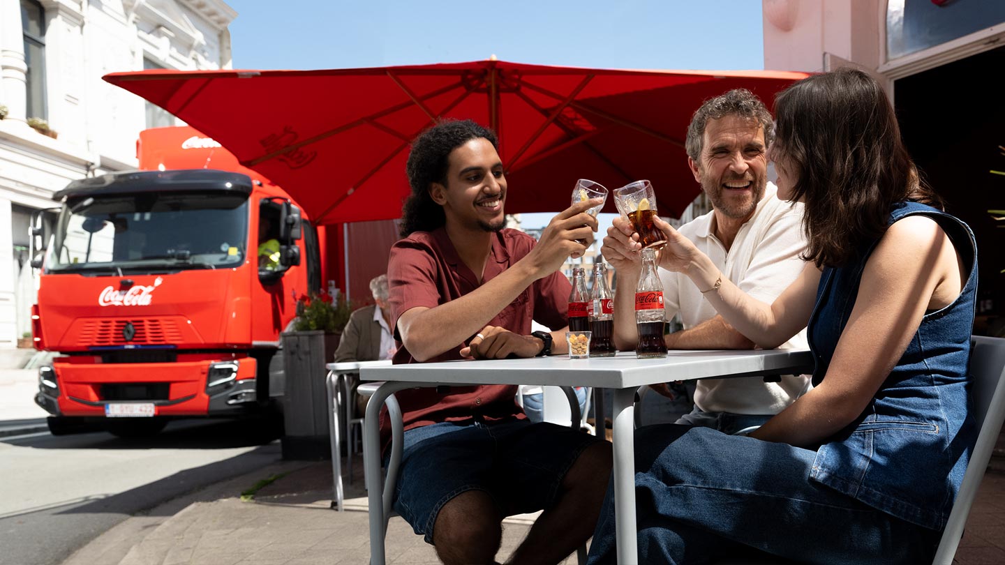 Drie personen zitten aan een tafel buiten en proosten met glazen Coca-Cola, terwijl flessen van het merk op de tafel staan. Op de achtergrond zijn een grote rode parasol en een rode vrachtwagen te zien in een zonnige straat.