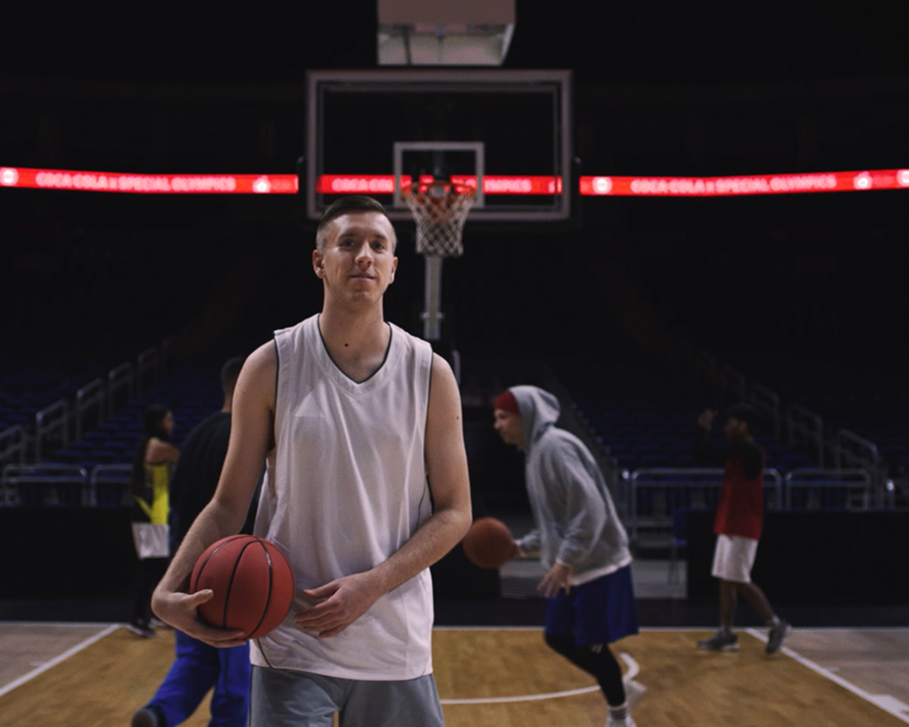 Een basketballer in een witte sportoutfit staat op een basketbalveld met een bal in zijn handen, terwijl op de achtergrond andere spelers actief zijn bij de basket.