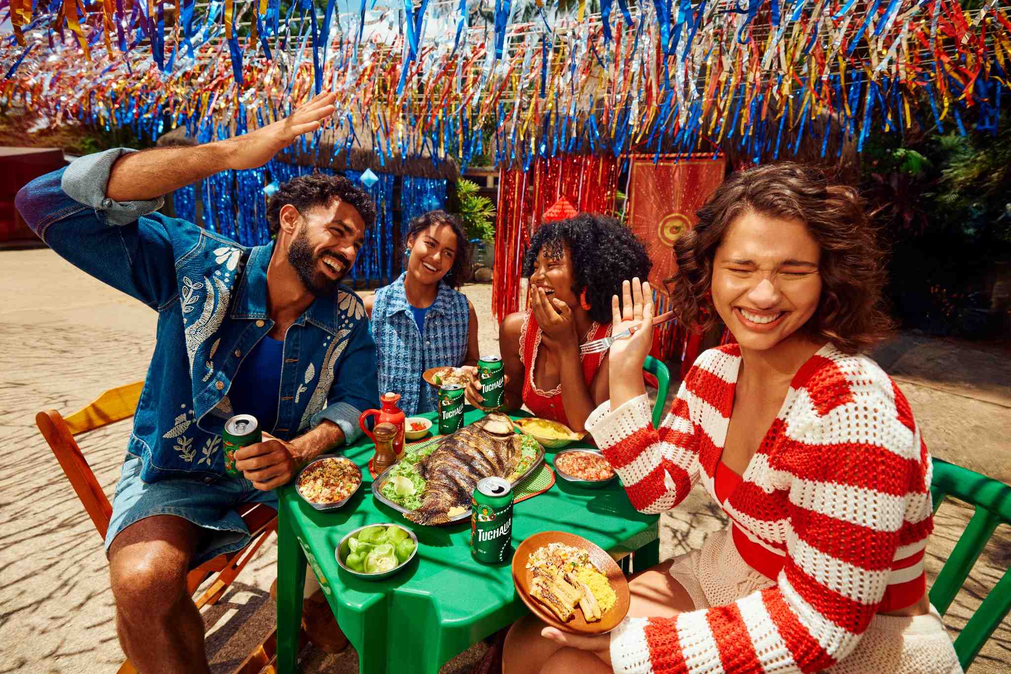 Quatro amigos sorrindo em um almoço festivo, com Tuchaua, peixe assado e outros pratos típicos em uma mesa verde, com fitas coloridas e decoração vibrante ao fundo.