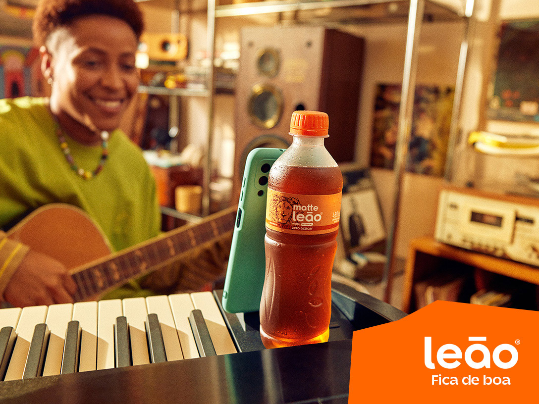 pessoa tocando violão enquanto olha para o celular e ele está apoiado em uma garrafa de Chas Leão em cima de um piano