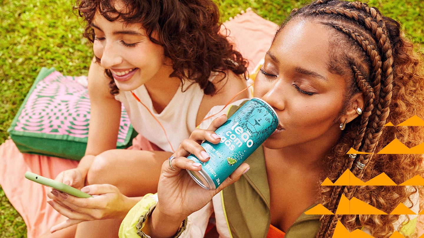 duas mulheres sentadas, uma delas sorri e usa o celular e a outra bebe um Chas Leão em lata.