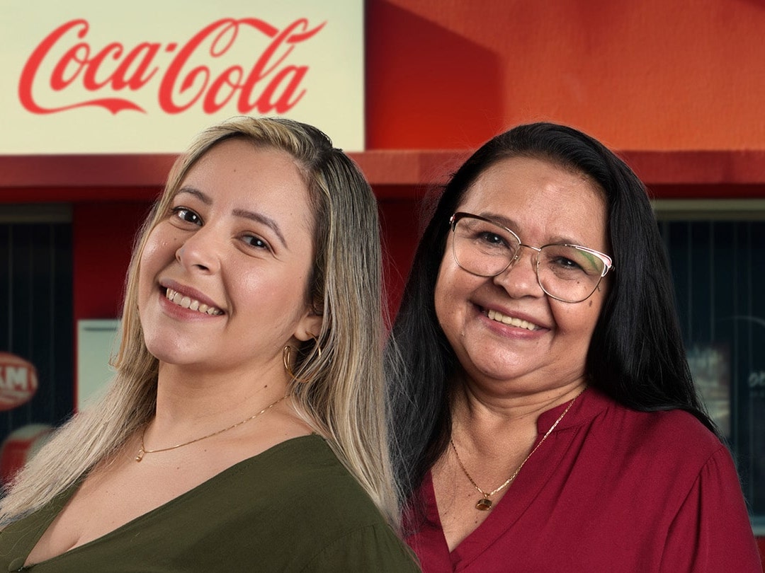 Duas mulheres sorrindo em frente a um letreiro da Coca-Cola, uma com cabelo loiro e outra com cabelo escuro e óculos.