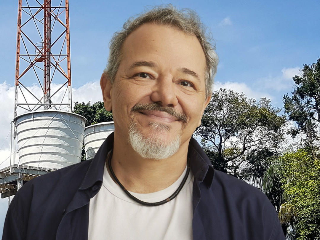 Um homem de meia-idade com barba e cabelo grisalho sorrindo, com uma torre de água e árvores ao fundo.