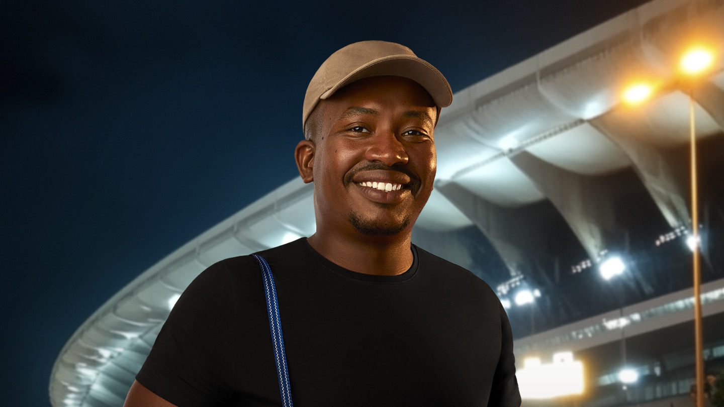 Homem sorridente de boné e camiseta preta, com estádio iluminado ao fundo.