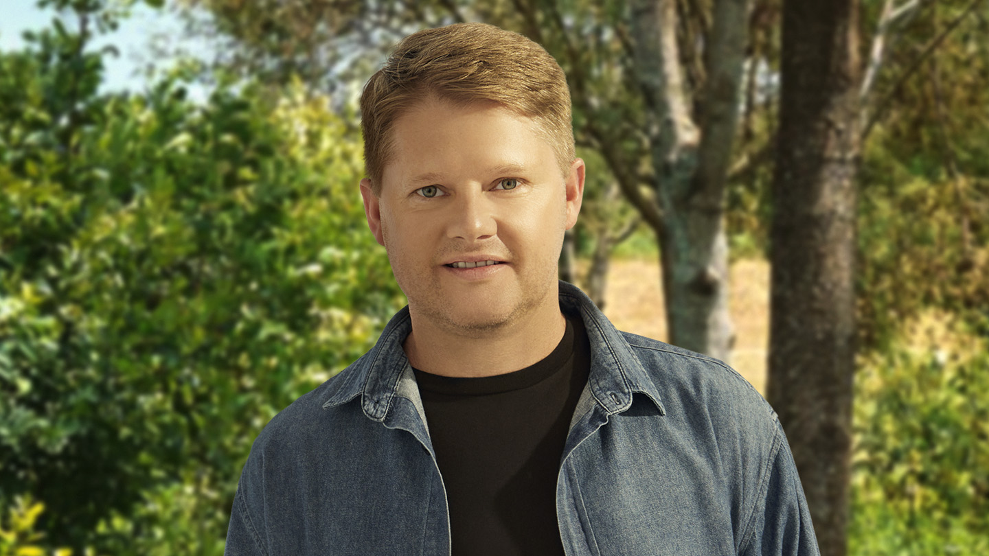 Homem loiro de camisa jeans e camiseta preta, em frente a árvores e folhagens.