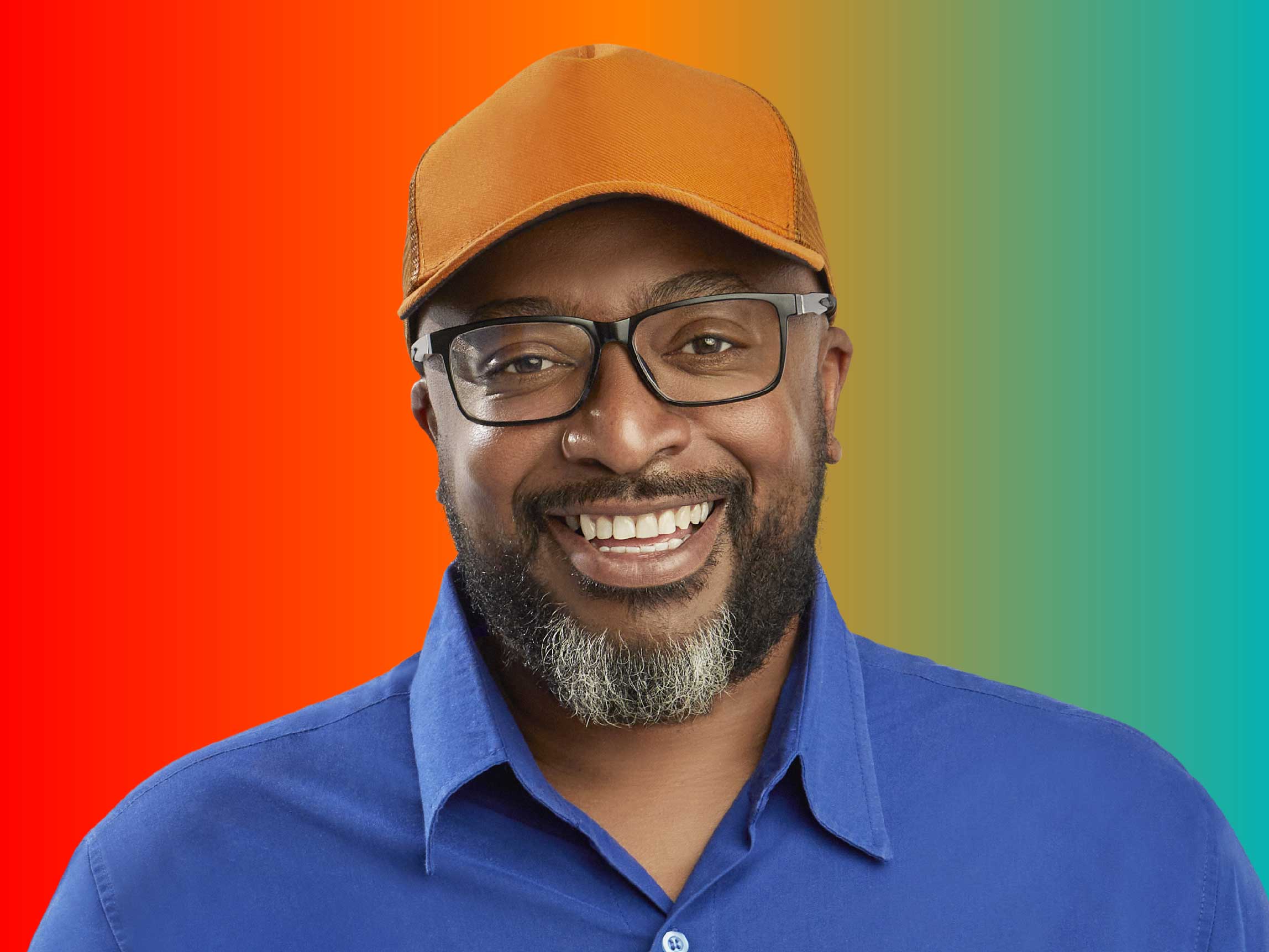 Homem sorridente de boné, óculos e barba, em fundo degradê laranja para azul.