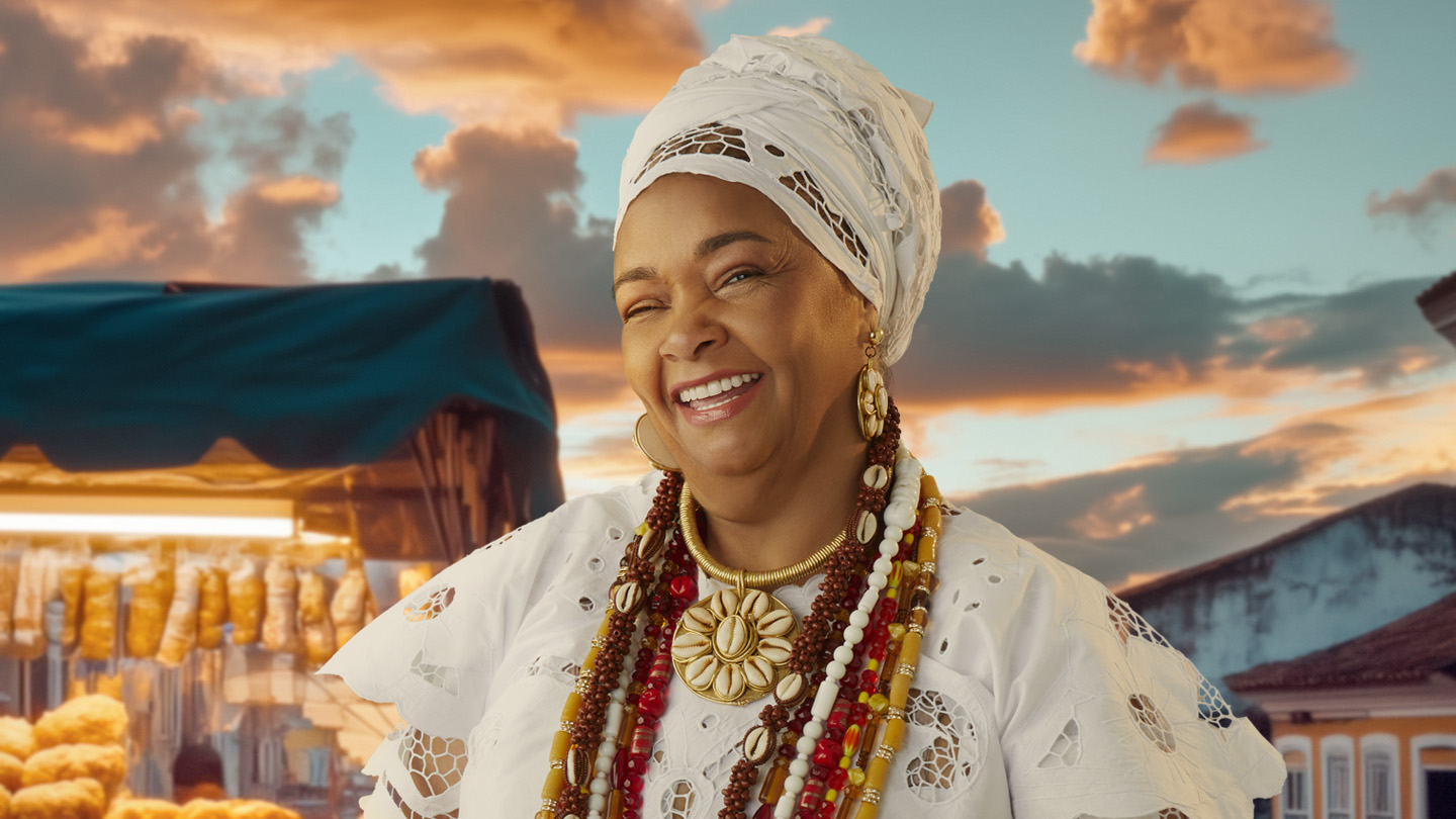 Mulher sorridente com turbante branco e colares coloridos, em frente a uma barraca de rua ao pôr do sol.