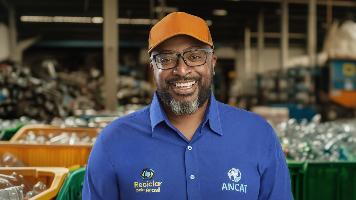 Homem sorridente de boné e camisa azul com logos de reciclagem, em um centro de coleta de materiais.