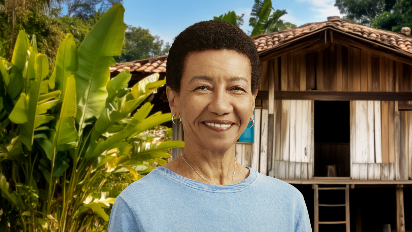 Mulher sorridente de cabelo curto e blusa azul, em frente a uma casa de madeira e folhagens.