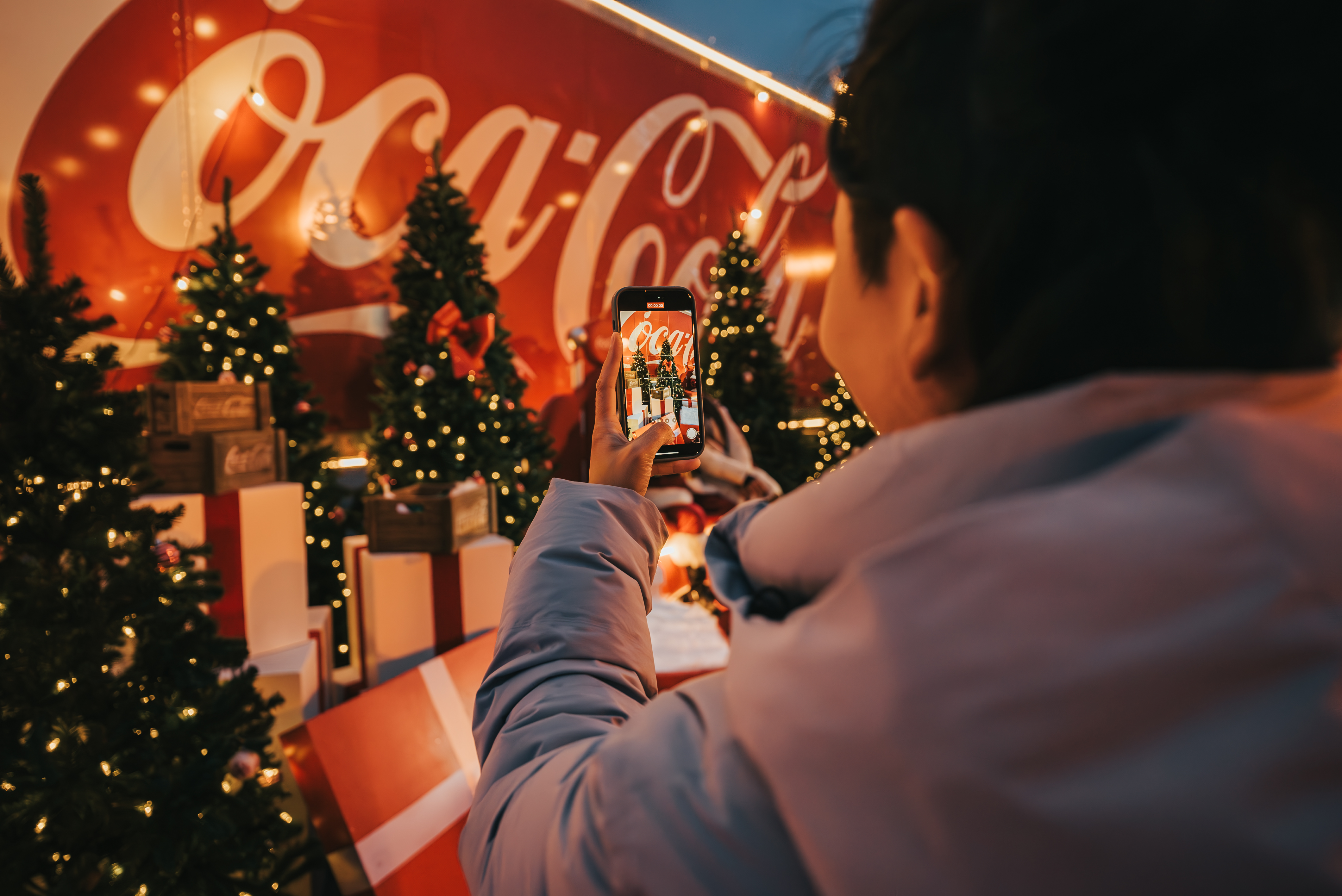 From over the shoulder, a person holds up a smartphone to photograph a festive scene with lit Christmas trees and presents in front of the red Coca-Cola Holiday Caravan truck.