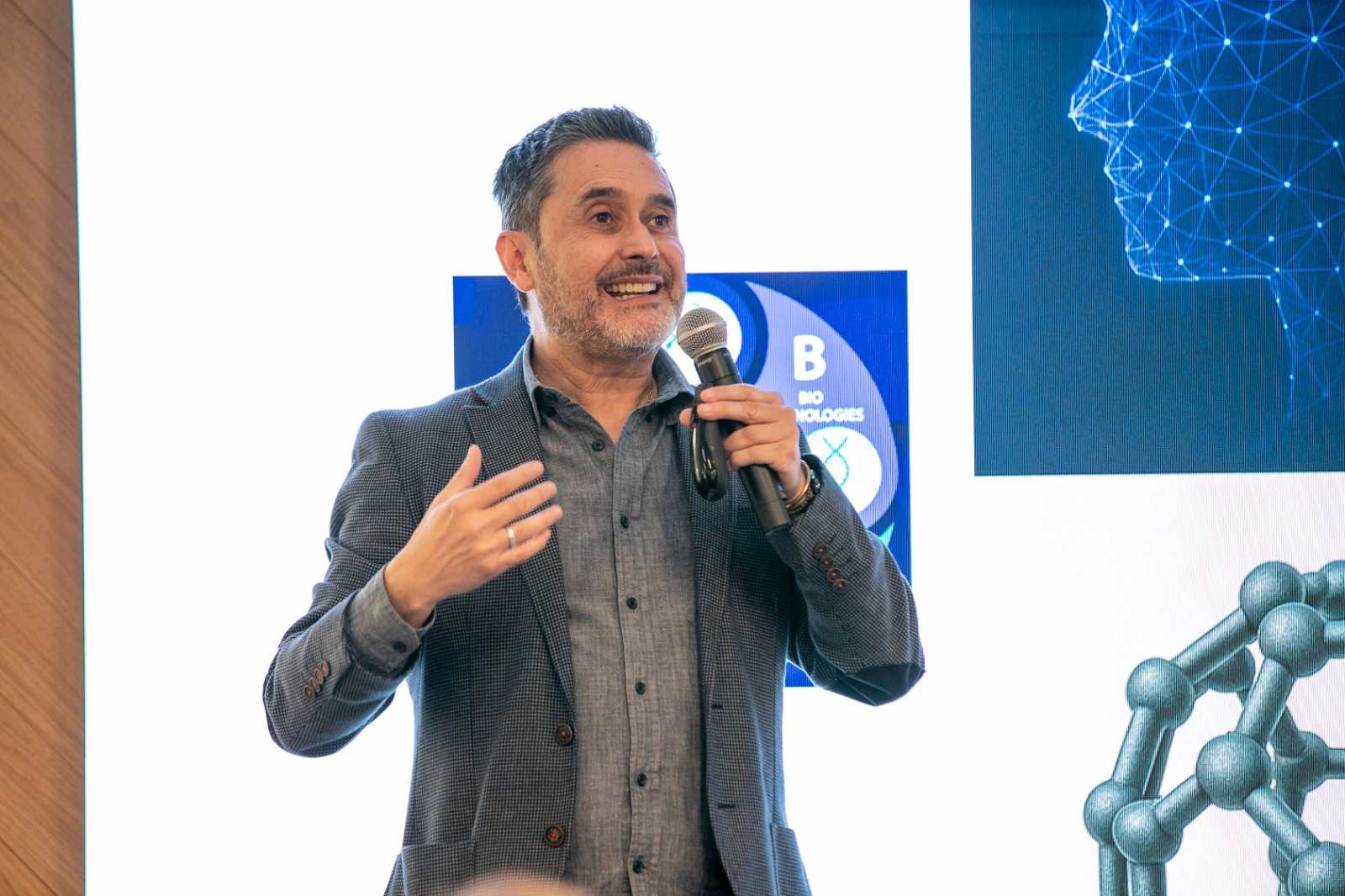 Hombre con un micrófono en la mano hablando con una pantalla detrás.