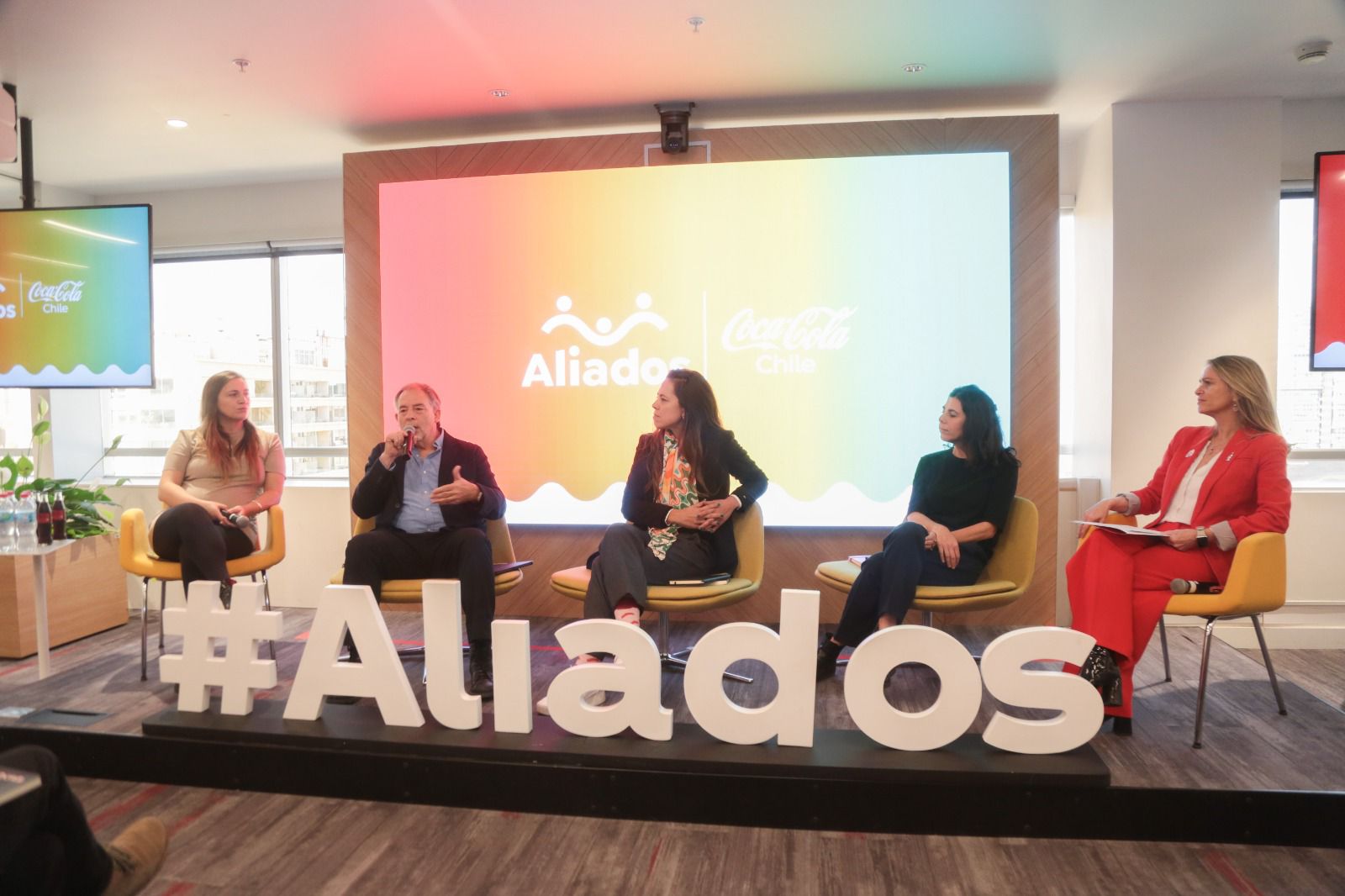 Grupo de personas charlando sobre una tarima. Enfrente de ellos, un cartel con las letras en blanco "Hashtag Aliados"