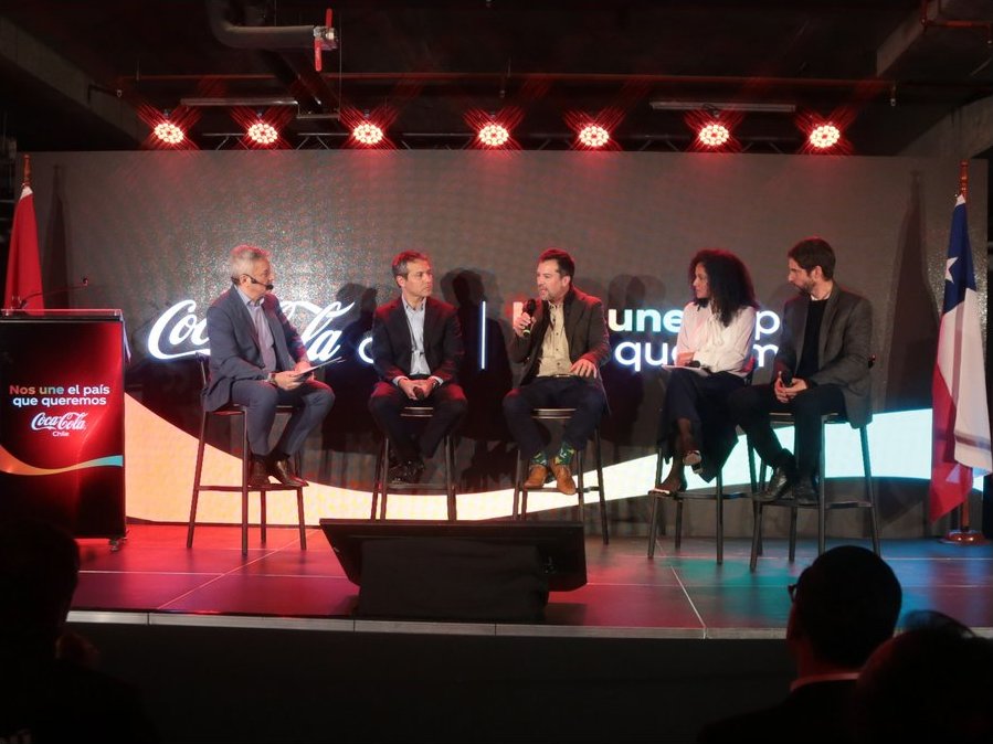 Un panel de discusión con cinco personas sentadas en taburetes altos en un escenario, con un telón de fondo oscuro que muestra el logo de Coca-Cola y el texto "Nos une el país que queremos".