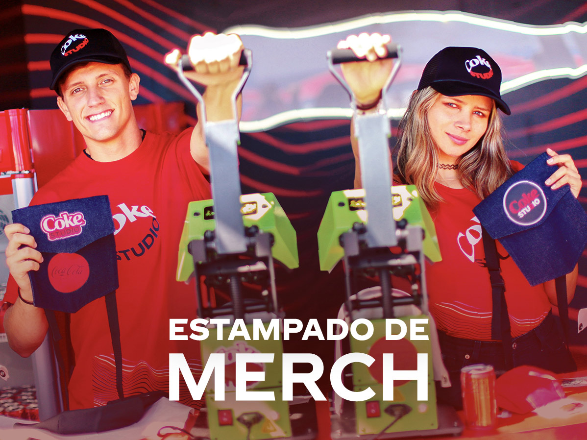 Dos personas en un stand de estampado de merchandising del festival Coke Studio, vistiendo camisetas rojas con el logo del evento. En el centro, se observan máquinas de estampado y productos personalizados como bolsos con diseños de Coke Studio. El texto "Estampado de Merch" aparece en la parte inferior, destacando la actividad creativa del lugar.
