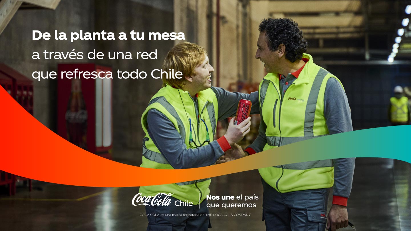 Dos colegas con chalecos de seguridad, uno rubio y otro moreno, conversando y riendo en un entorno industrial. Uno sostiene una lata de Coca-Cola.