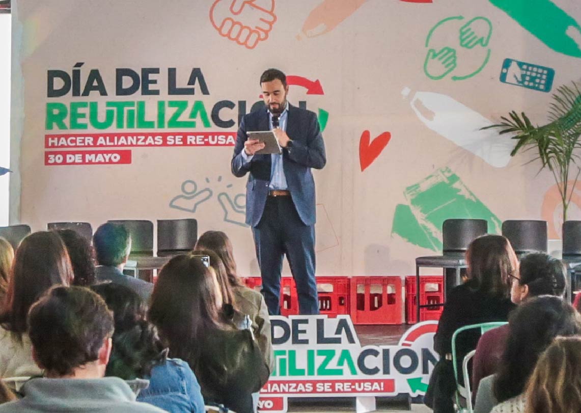 Hombre dando charla sobre reutilización en evento, con público atento y mural temático de fondo.