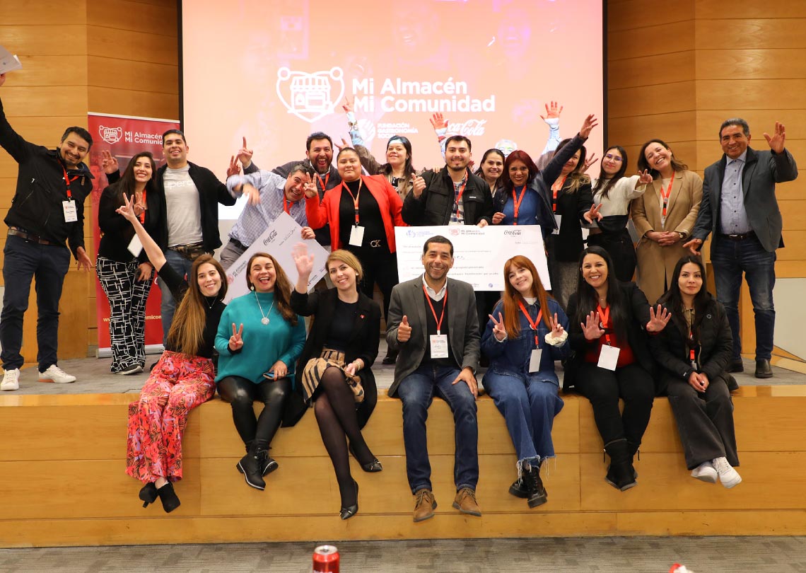 Un grupo diverso celebra con entusiasmo en un evento, posando frente a un cartel de "Mi Almacén Mi Comunidad" y Coca-Cola, con un cheque gigante.