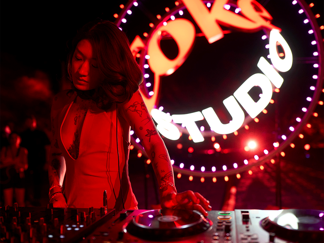 Peggy Gou tocando en vivo en un evento de Coke Studio, con iluminación vibrante en rojo y el logo al fondo. Lleva un vestido rojo y tiene tatuajes visibles en los brazos. Al fondo, hay personas y un ambiente de fiesta y música