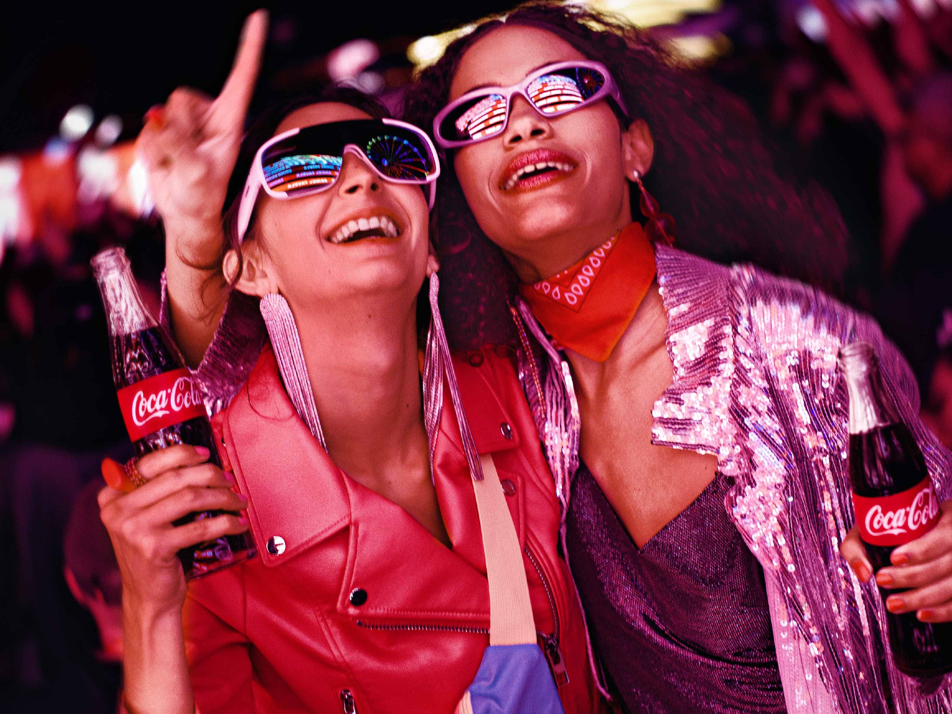 Dos mujeres usando gafas en un ambiente festivo nocturno disfrutando de una Coca-Cola.