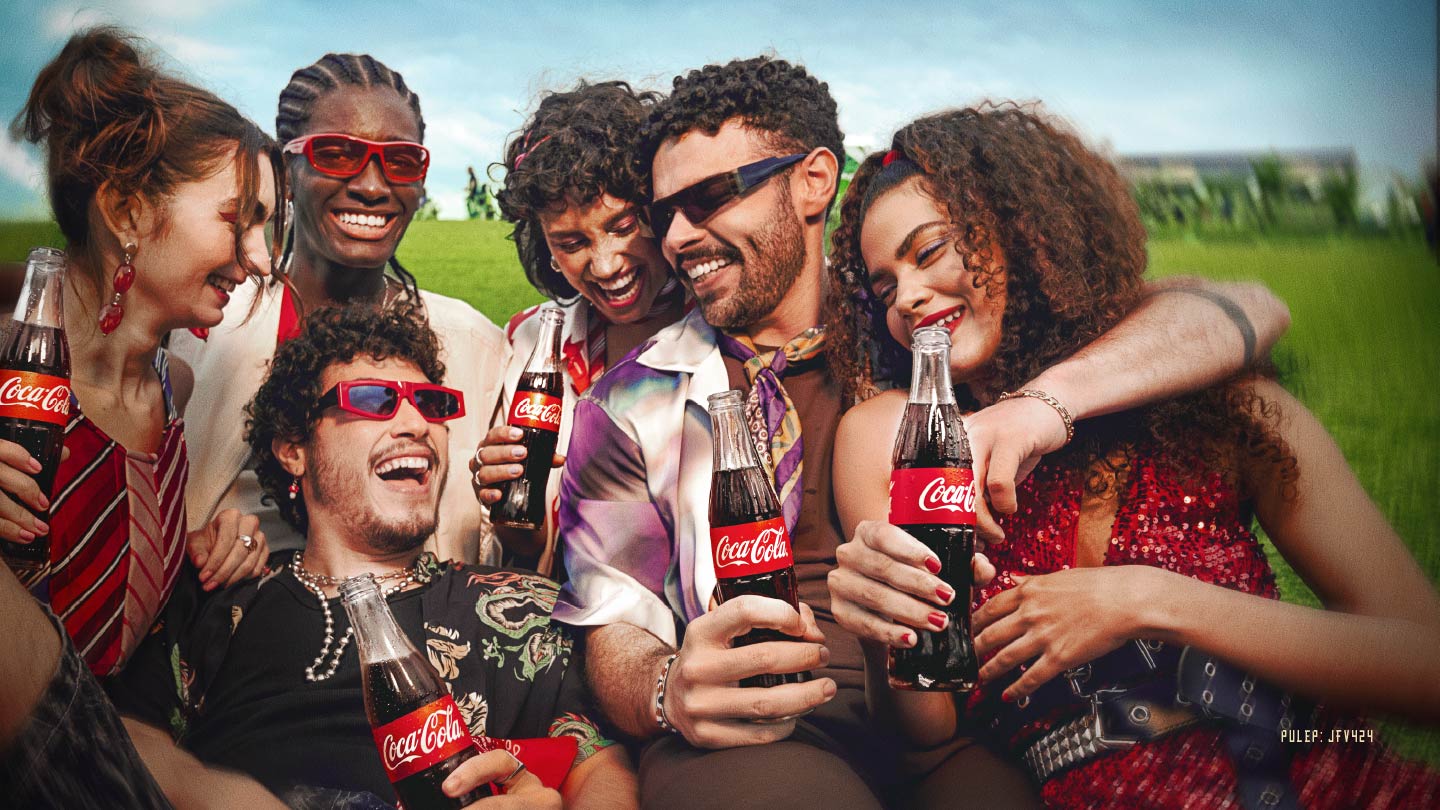 Un grupo de seis jóvenes diversos, sonrientes y alegres, posan juntos al aire libre, sosteniendo botellas de Coca-Cola. La escena sugiere un ambiente festivo y de disfrute.