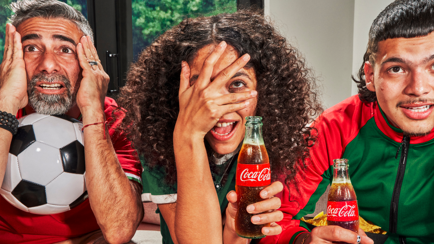 Drie voetbalfans in rood-groene shirts juichen met opgeheven vuisten; een sjaal wordt omhooggehouden en een voetbal is zichtbaar op de achtergrond.