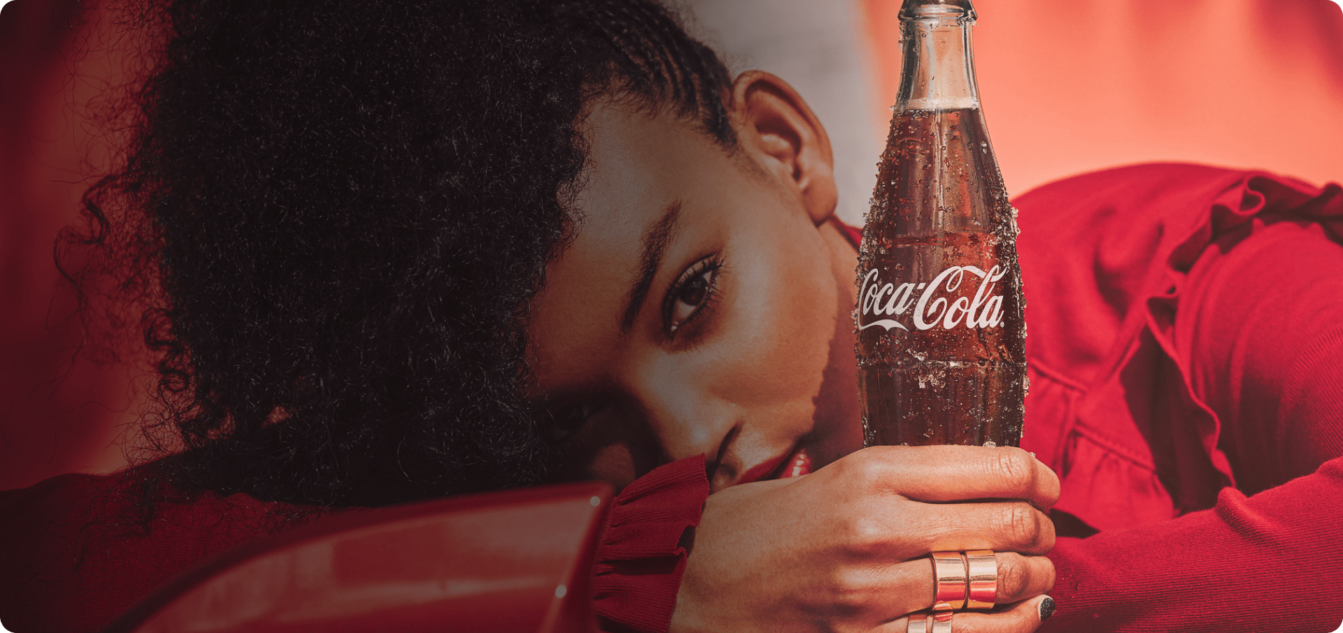 Dorë mban një shishe Coca-Cola me pika të ftohta, në sfond veshje të kuqe.