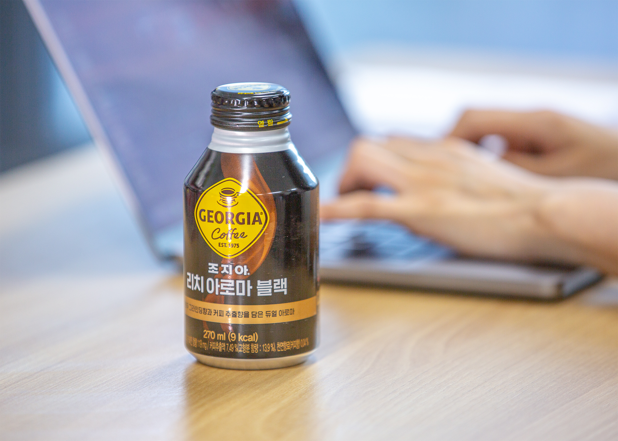 책상 위에 놓인 조지아 리치 아로마 블랙 제품에 초점이 맞춰져 있고, 제품 뒤로는 노트북 키보드를 누르는 두 손의 형체를 확인할 수 있다.