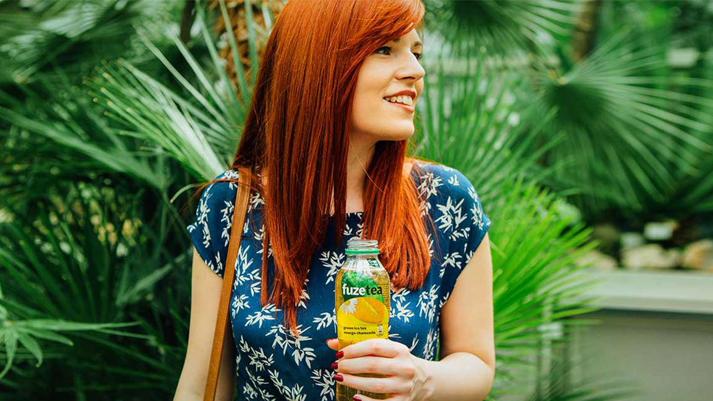 Grua me flokë të kuqe duke qeshur, mban një shishe Fuze Tea me shije mango-kamomil, në sfond gjethe jeshile.