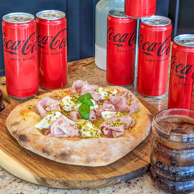 Picë me proshutë, mocarela dhe fistikë, e rrethuar nga kanaçe Coca-Cola Zero Sugar.