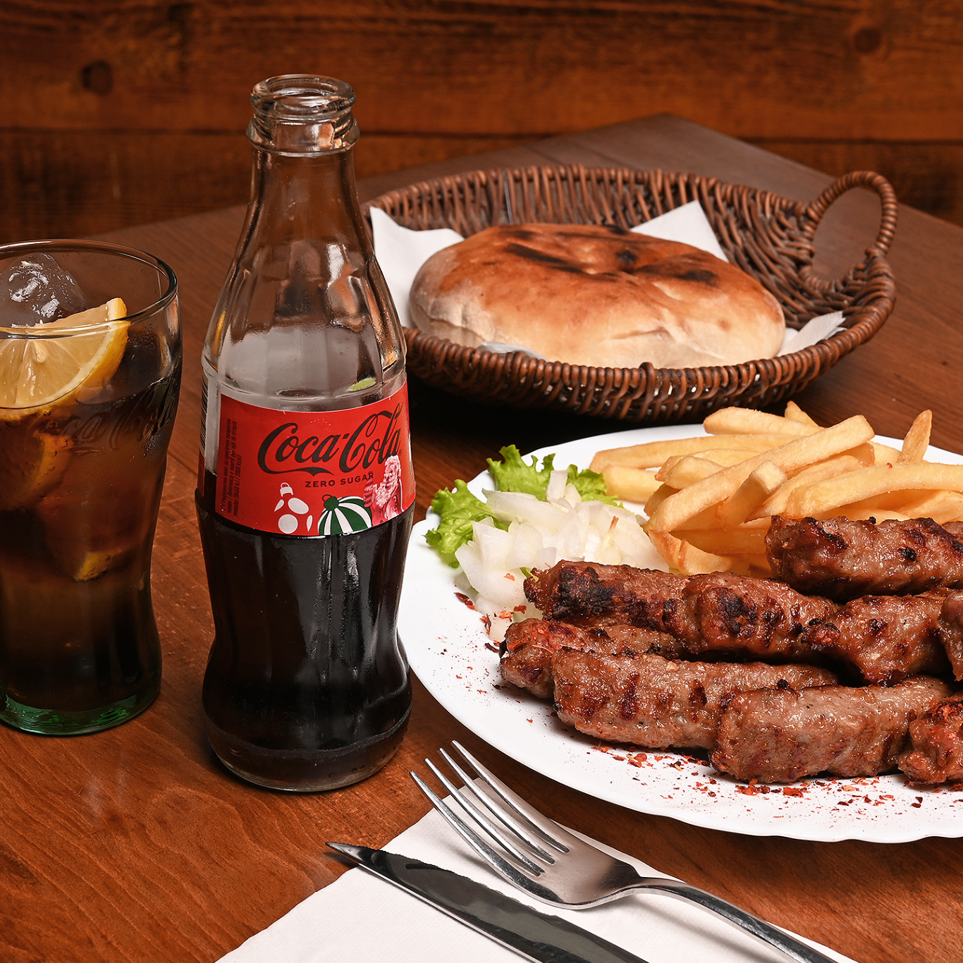 Pjatë me qebapë, patate të skuqura, bukë dhe një gotë Coca-Cola Zero.
