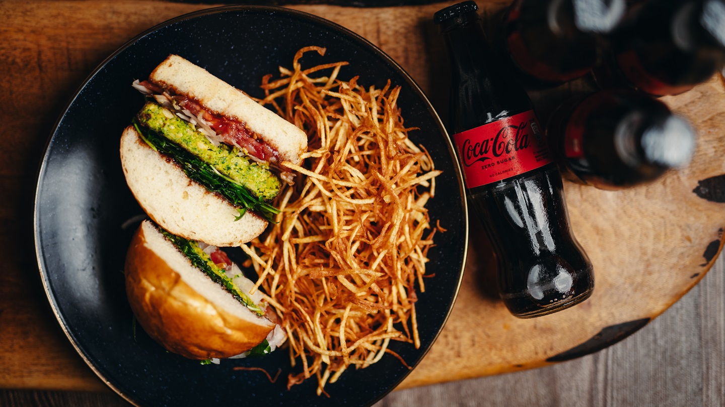 Gjysma burgeri me patate të skuqura dhe një shishe Coca-Cola Zero Sugar në një tavolinë druri.