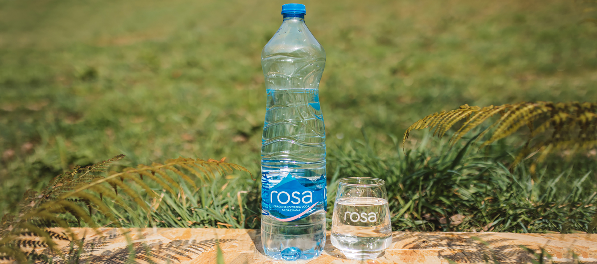 Shishe plastike dhe gotë uji Rosa në një sipërfaqe druri me bar të gjelbër në sfond.