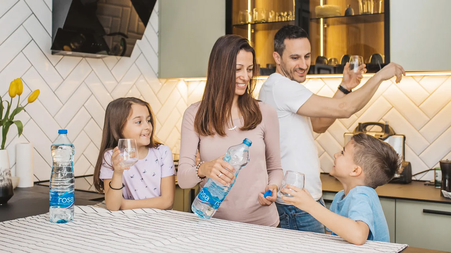 Familje në kuzhinë me shishe uji Rosa dhe gota në tavolinë.
