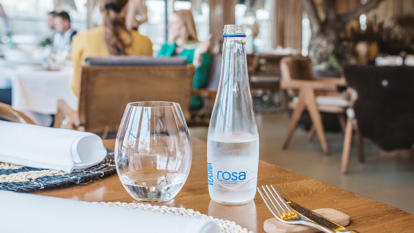 Shishe dhe gotë uji Rosa në tavolinë restoranti me takëm të shtruar.