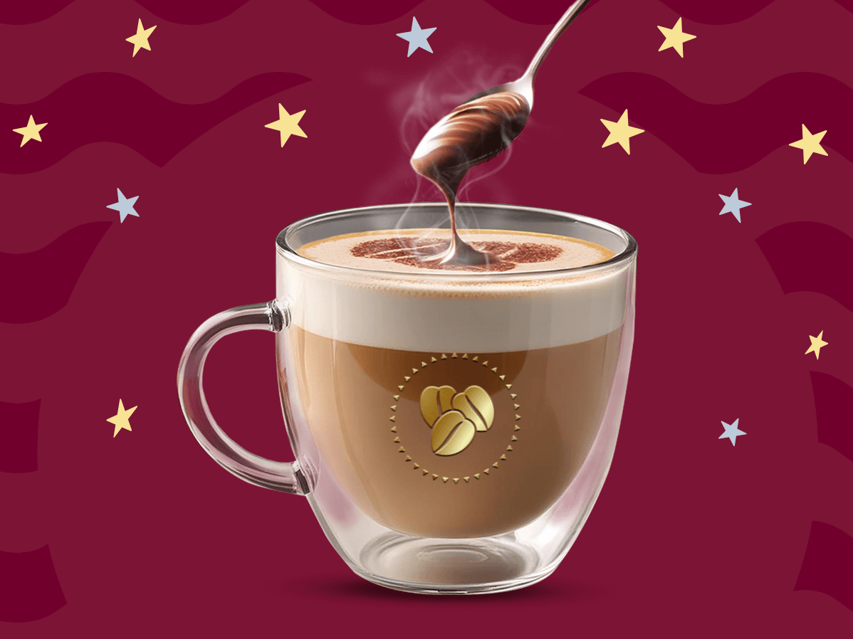 Una taza de cristal con café latte caliente y una cuchara por encima agregando crema de avellanas sobre un fondo en tonos guinda.