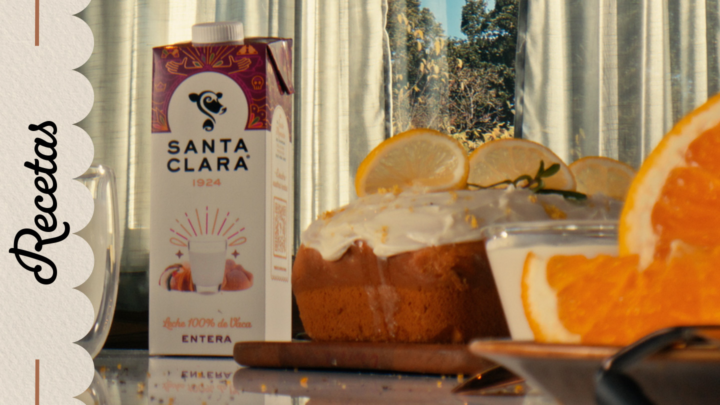 Leche entera Santa Clara junto a un pastel glaseado con rodajas de limón y un vaso con leche y rodaja de naranja, en una mesa iluminada por la luz natural.  