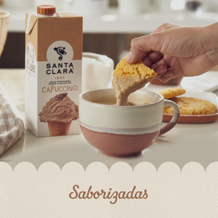 Leche sabor cappuccino Santa Clara acompañada de una taza con leche y una mano sumergiendo una galleta. Al fondo, más galletas sobre un plato.